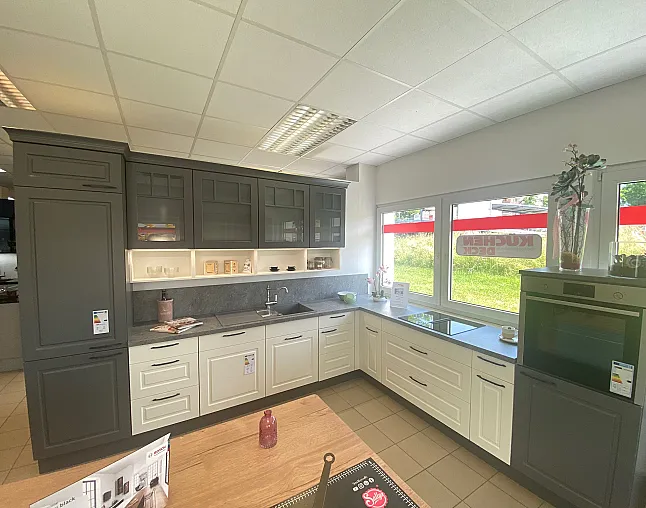 Moderne U-förmige Küchenzeile mit grauen und weißen Schränken, dunkler Arbeitsplatte, eingebautem Herd und großem Fenster mit Blick auf eine grüne Wiese.