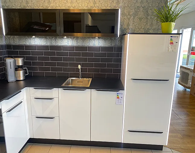 Moderne Küchenzeile mit weißen Schränken, schwarzer Arbeitsplatte, Edelstahlspüle, Kaffeemaschine und großem weißen Kühlschrank mit Pflanze oben.